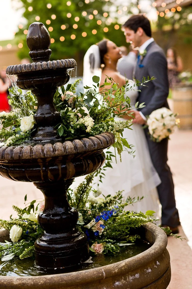 A romantic wedding scene with a couple in focus behind a flower-adorned fountain. Soft lights hang above, creating a warm, celebratory atmosphere during a Villa Parker wedding in Parker, Colorado.