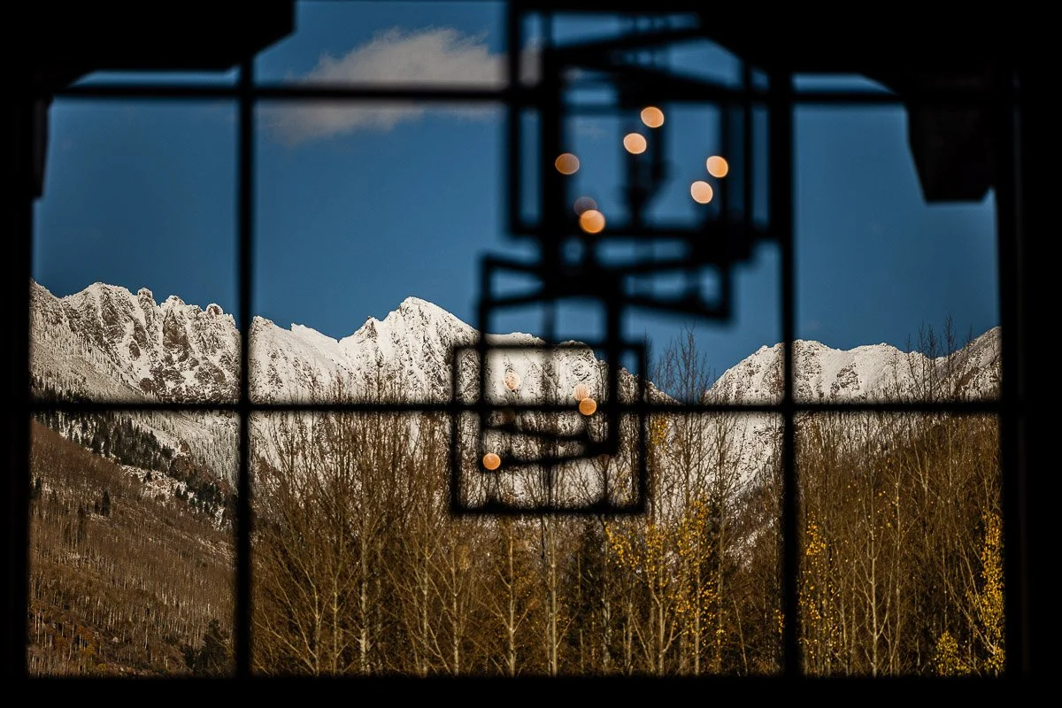 A snow-capped mountain is framed by a window, with a geometric chandelier in the foreground. The blue sky and bare trees create a peaceful, wintry scene.
