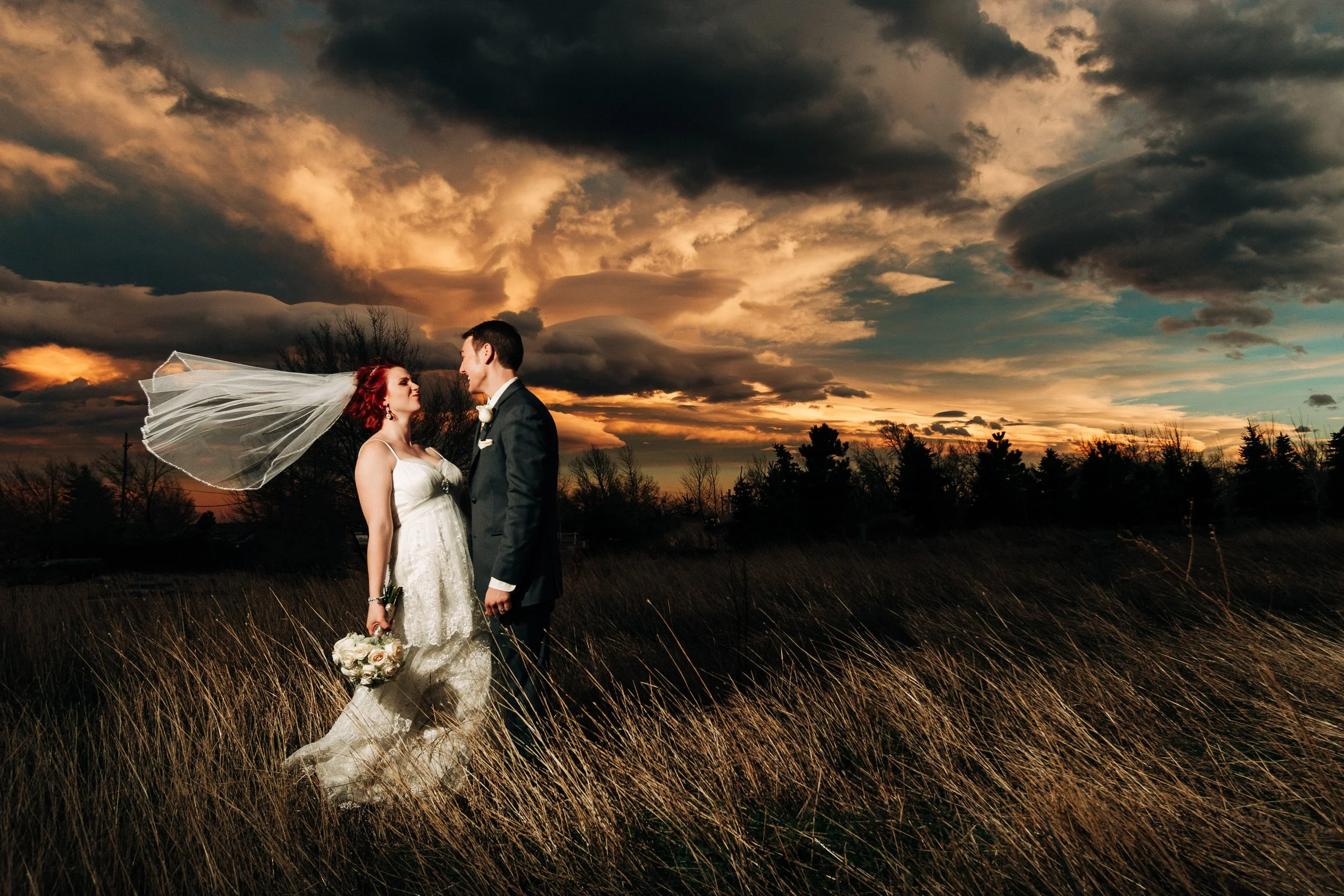 A bride's veil blows in the wind as she jokes with her groom in front of a dramatic winter sunset during a Greenbriar Inn wedding reception in Boulder, Colorado