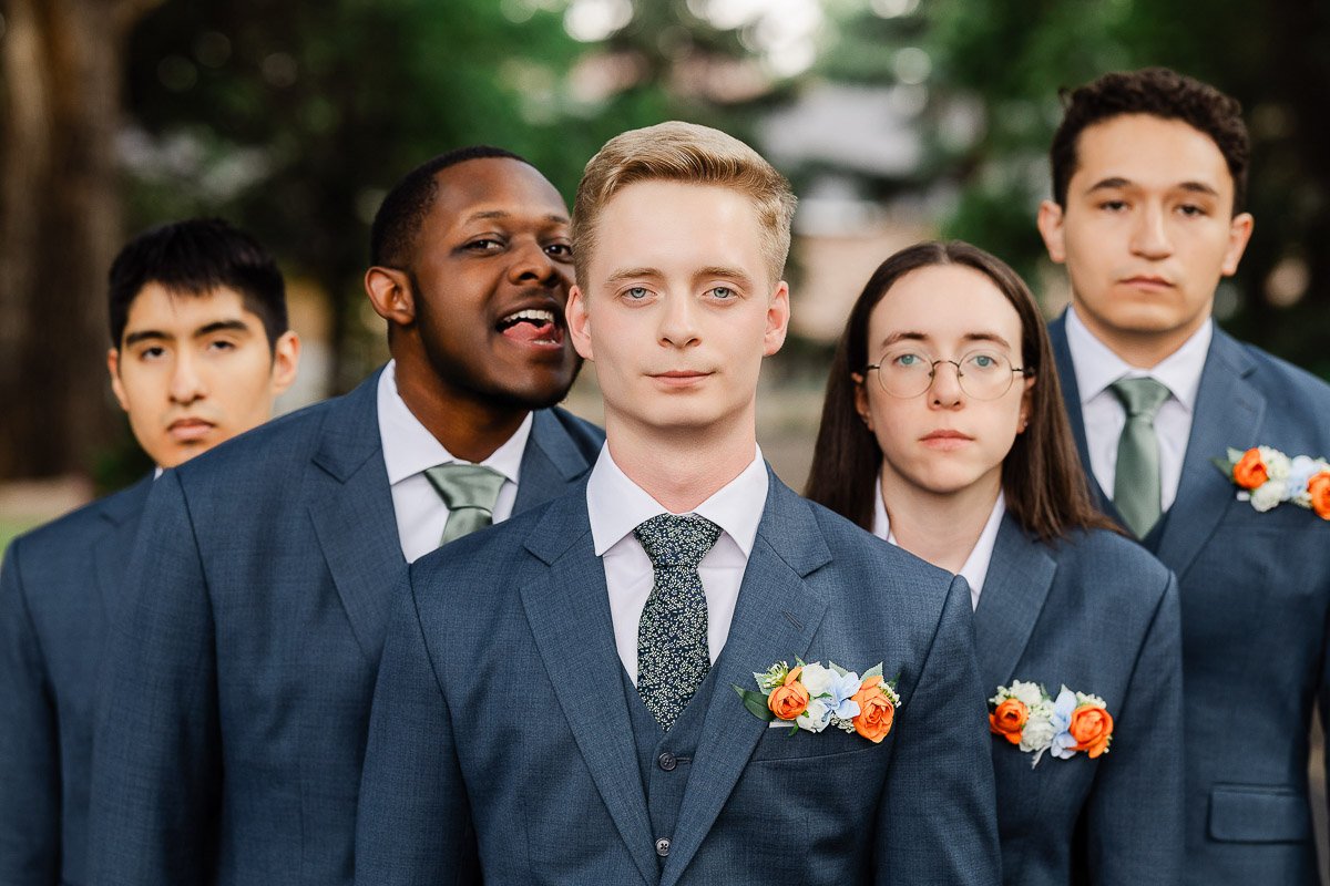 A group of five men wearing blue suits with orange boutonnieres pose outdoors. They display various expressions, from playful to serious.