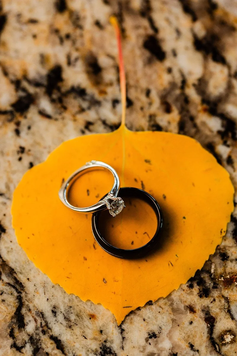 A diamond engagement ring and a black wedding band rest on a vibrant, heart-shaped yellow leaf, set against a textured stone background.