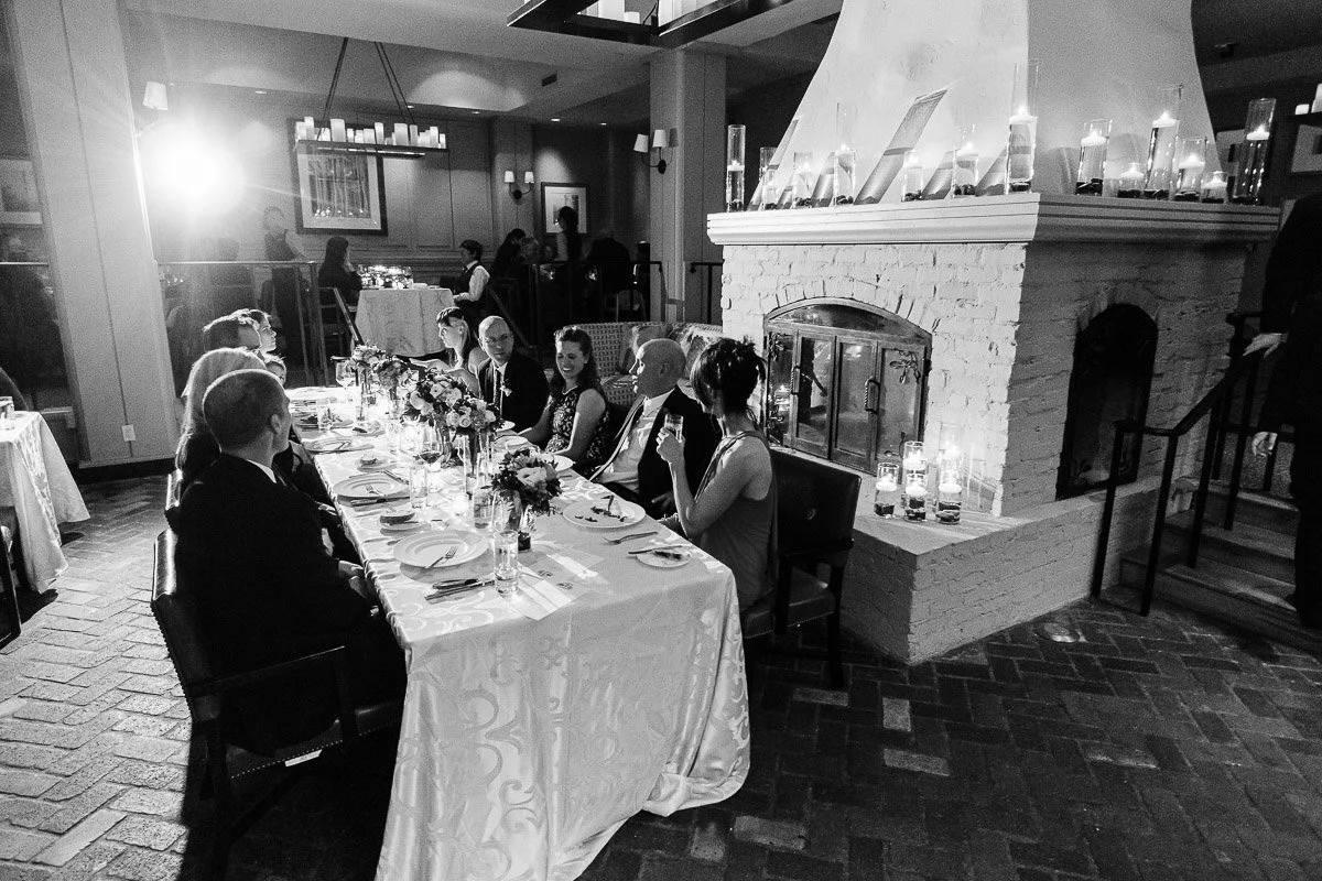 Black-and-white image of a formal dinner with ten people seated at a long table near a fireplace. Candles and flowers create an elegant ambiance during a Park Hyatt wedding reception in Beaver Creek, Colorado.