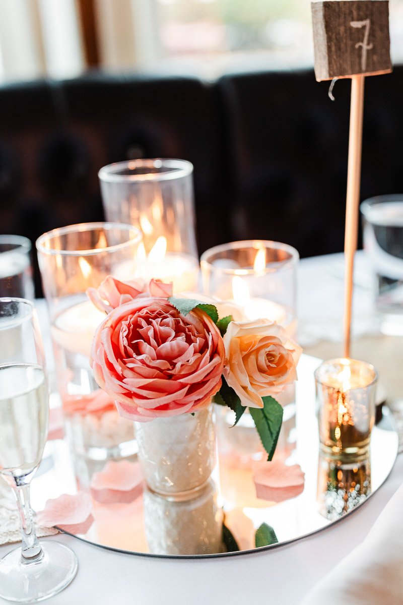 Elegant table setting with lit candles and a centerpiece of pink and peach roses on a mirrored base. Champagne glass nearby. Romantic ambiance.