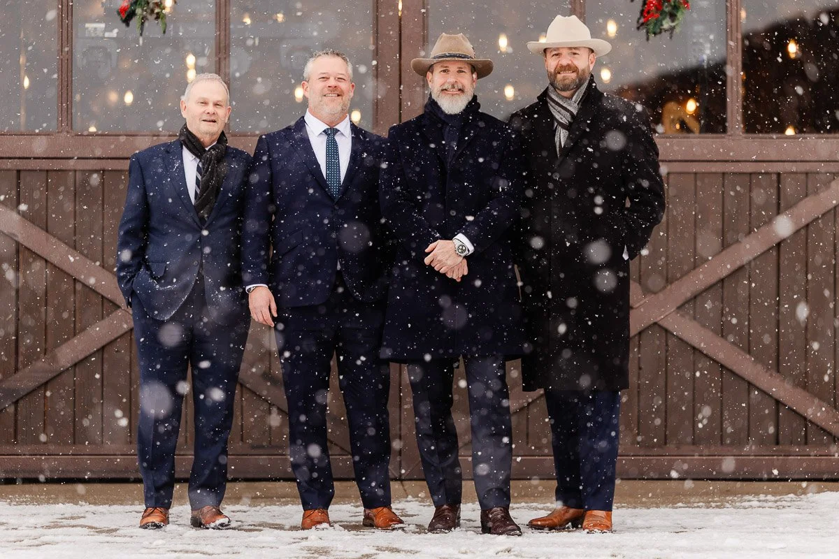 Four men in suits and coats stand smiling in front of a rustic barn door amidst falling snowflakes. The mood is festive and warm despite the wintry setting.