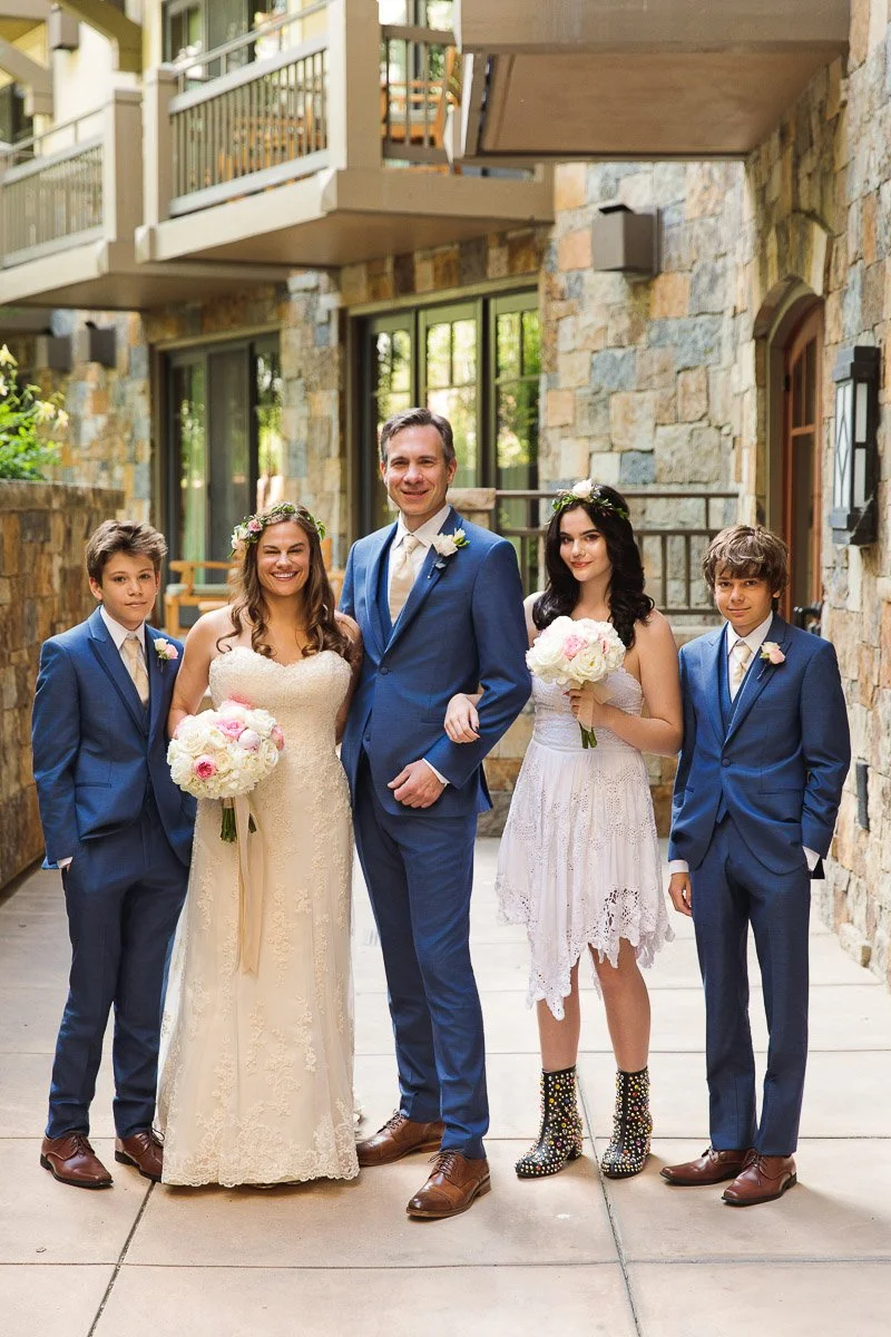 Wedding party photo in a stone-paved corridor. A couple in formal attire is flanked by three teenagers, all smiling. The mood is joyful and elegant.