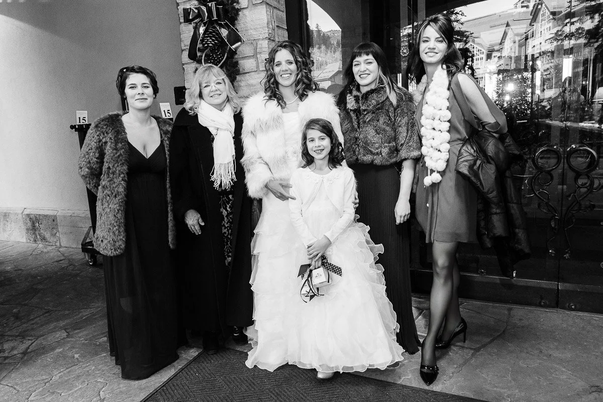 A group of six people poses happily outside, dressed elegantly for a formal event. A young girl in a white dress stands in front, smiling.