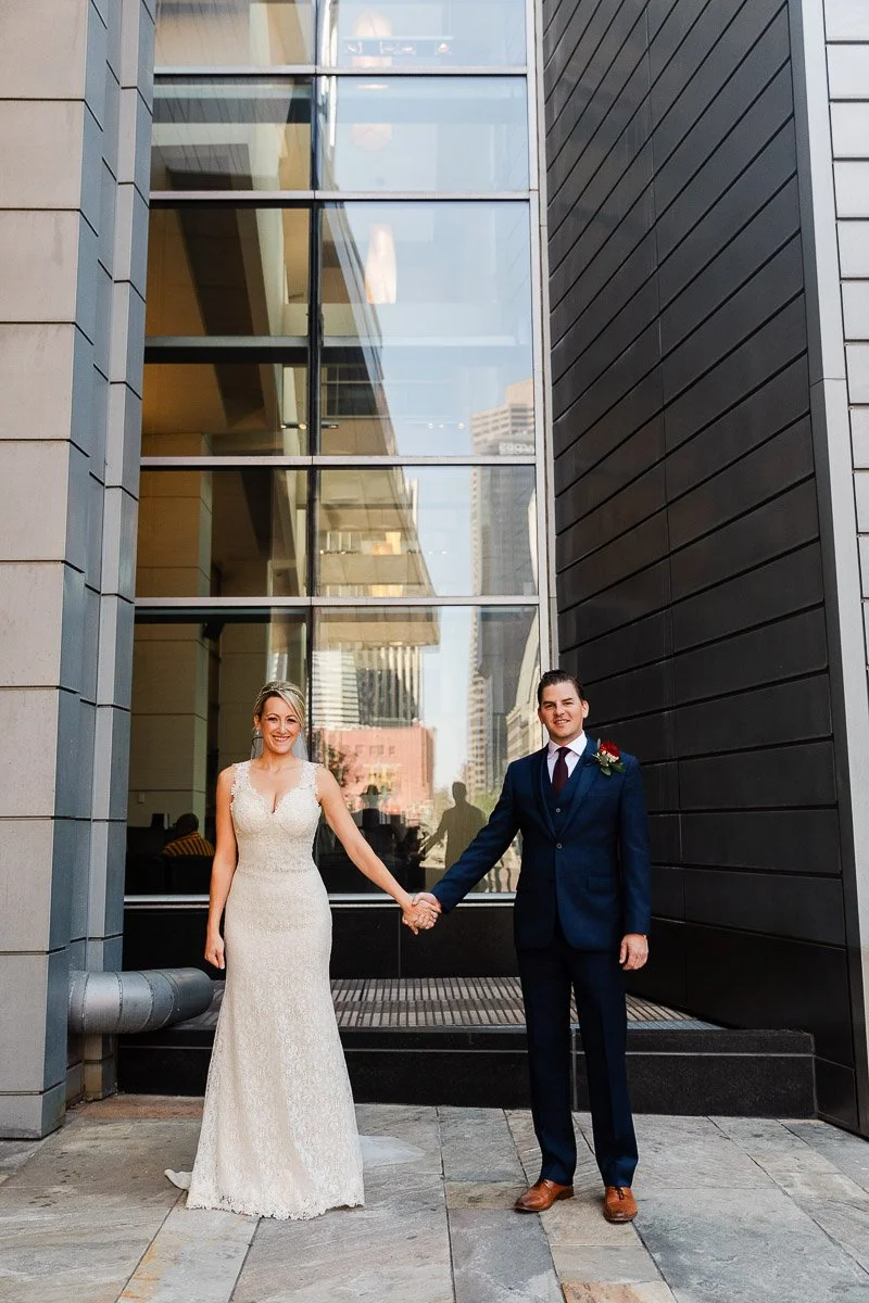 A bride in a lace gown and a groom in a navy suit stand hand in hand, smiling in an urban setting with reflective glass and stone walls during a Coohills Restaurant wedding in Denver, Colorado