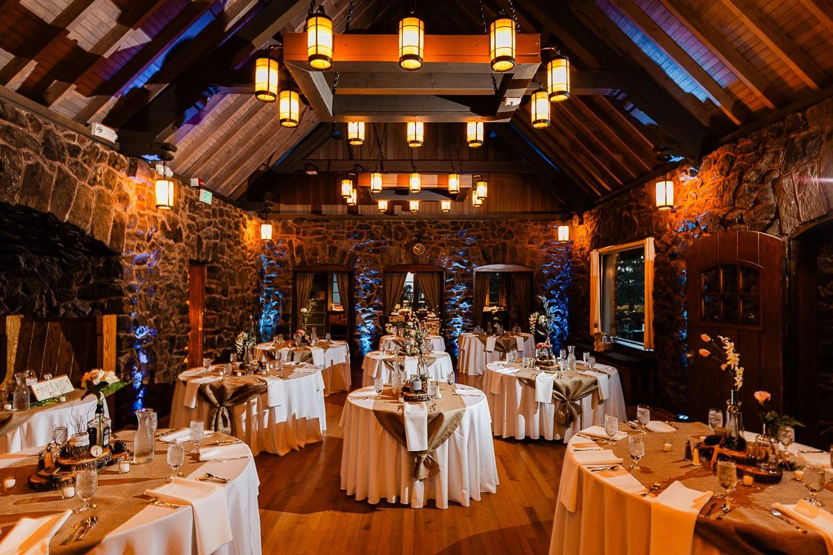 Elegant dining hall with rustic stone walls and wooden beams, warmly lit by hanging lanterns. Round tables are dressed in white and burlap, creating a cozy, romantic atmosphere.