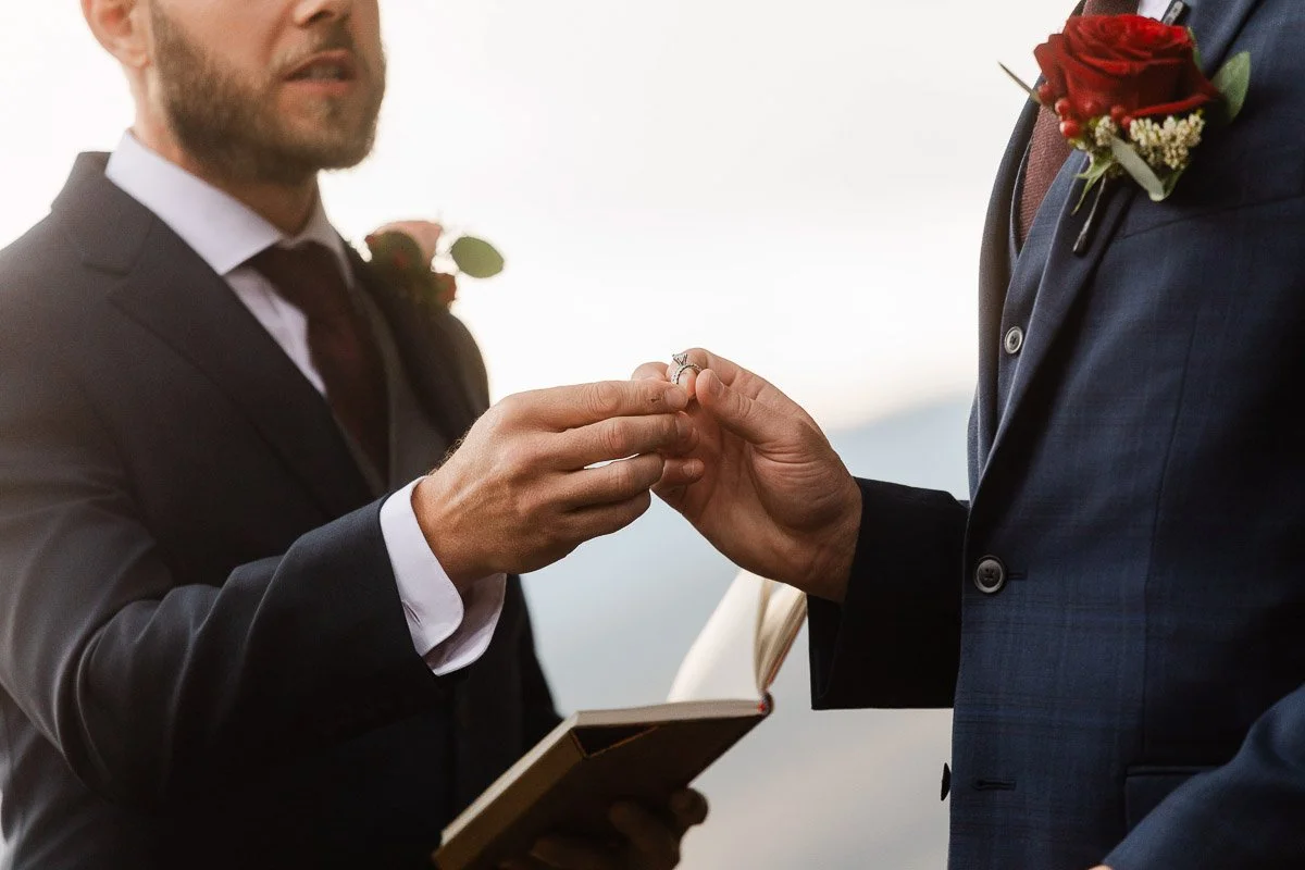 Two men in suits exchange rings during a wedding ceremony. They wear red rose boutonnieres, symbolizing love and commitment. The mood is intimate and joyful.