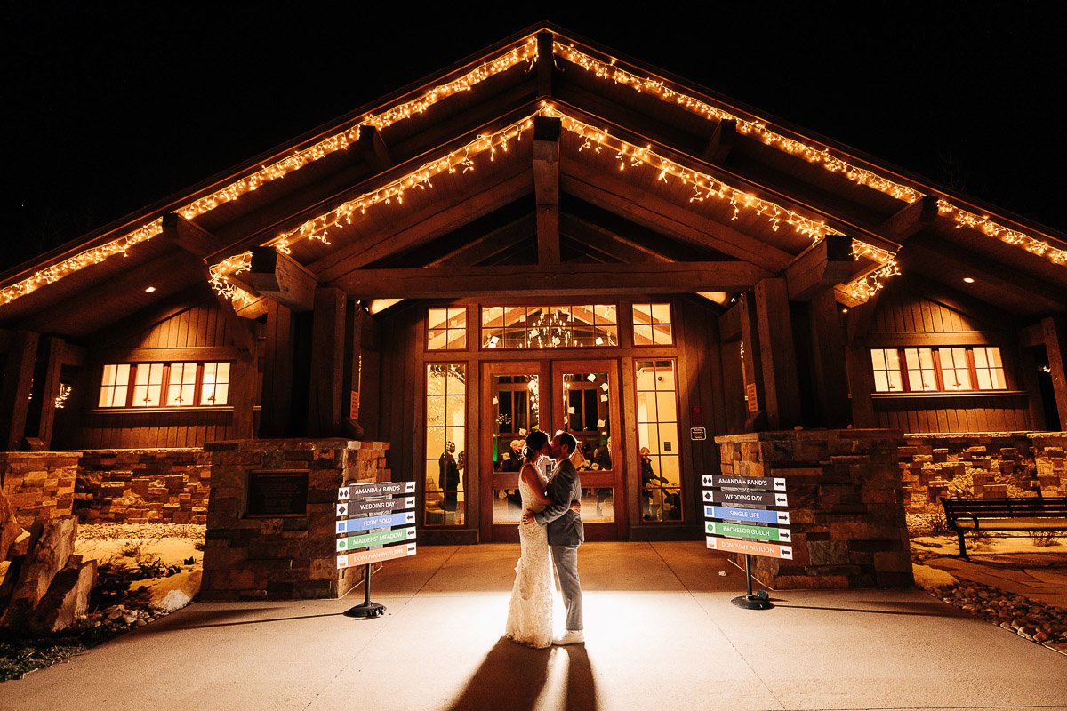 A couple, illuminated by soft lighting, embraces in front of a warmly lit rustic lodge — Donovan Pavillion — decorated with twinkling string lights on a clear night, captured by Vail wedding photographer, tomKphoto