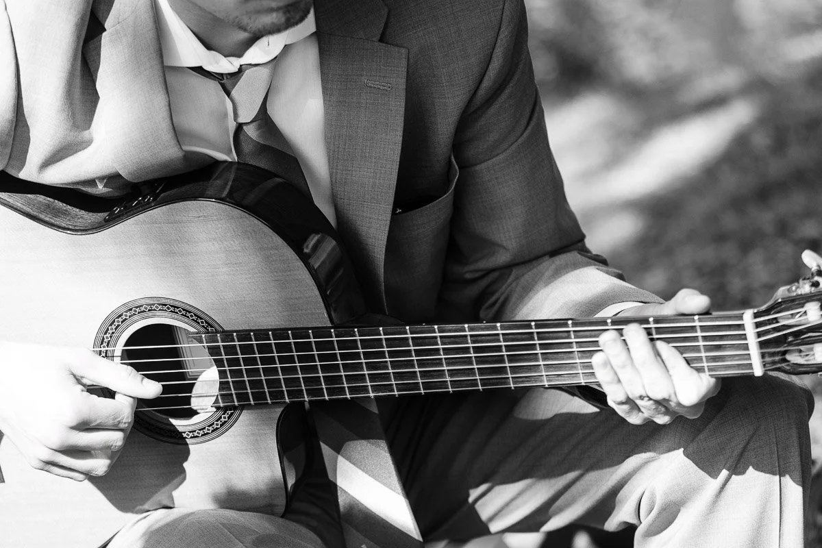 A man in a suit plays an acoustic guitar in a sunlit outdoor setting. His head is slightly bowed, creating a calm, contemplative mood. Black and white.