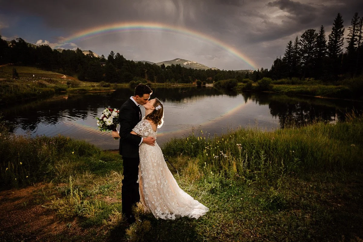 A couple kisses by a serene lake under a vibrant rainbow. The bride in a lace gown holds a bouquet. Dark clouds and lush greenery set a romantic mood caputred by Breckenridge wedding photographer tomKphoto
