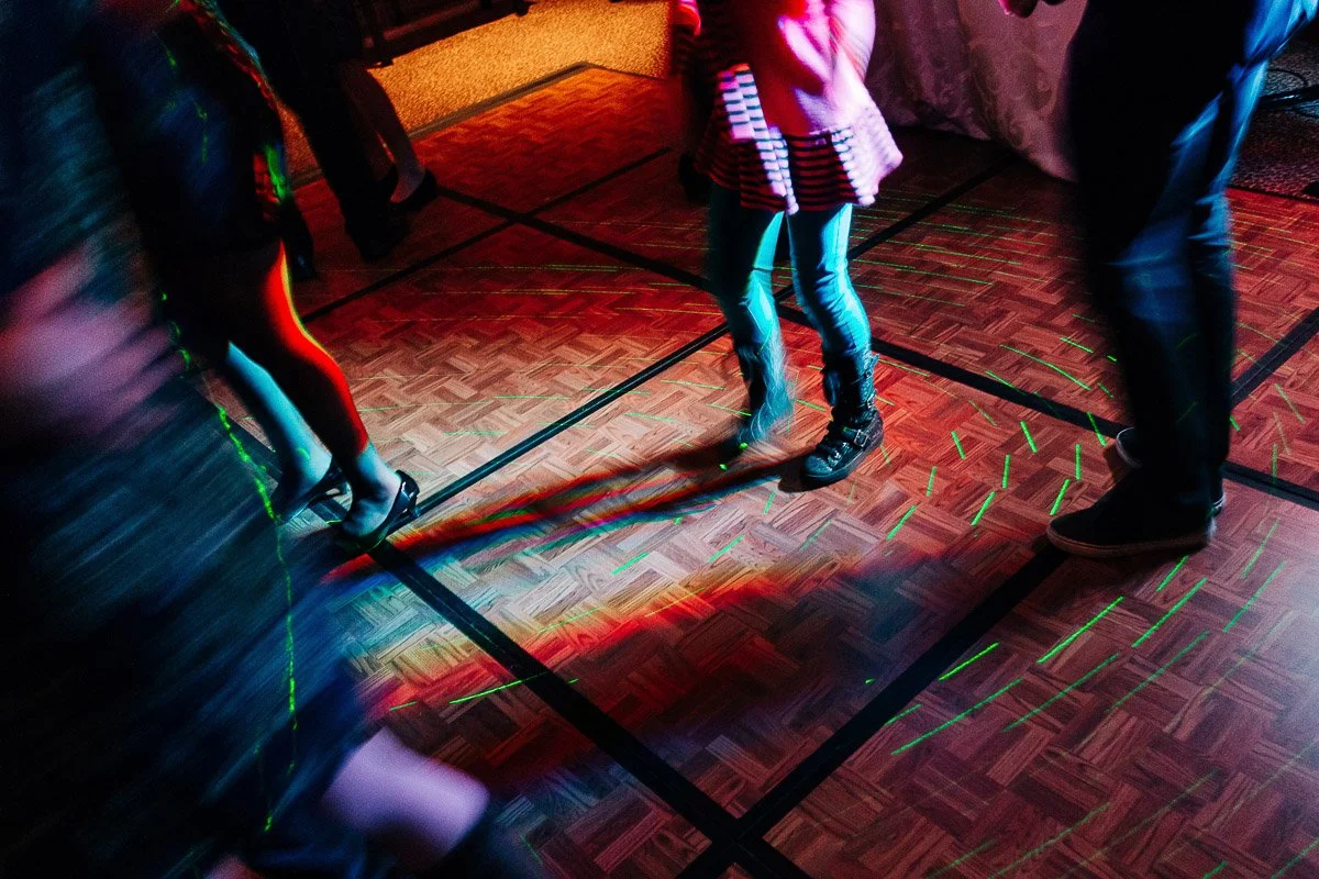Blurred image of people dancing on a wooden floor, bathed in colorful lights. The scene conveys energy and movement, suggesting a lively party atmosphere.