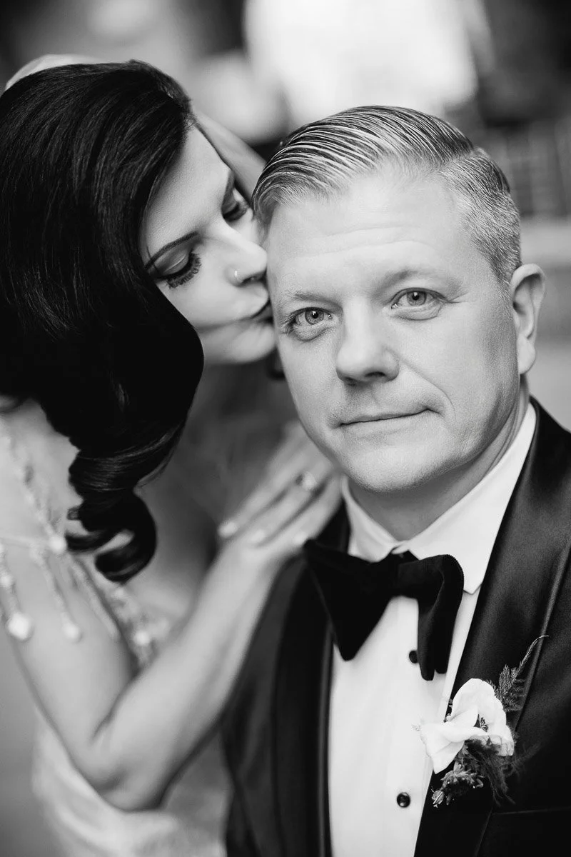 Black and white portrait of a woman kissing a man's temple. He wears a tuxedo with a boutonniere, exuding elegance and tenderness.