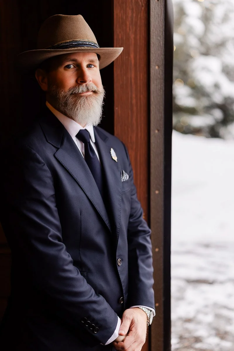 Man with a gray beard in a hat and navy suit stands confidently in a wooden doorway. Snowy background suggests a cold, serene setting.