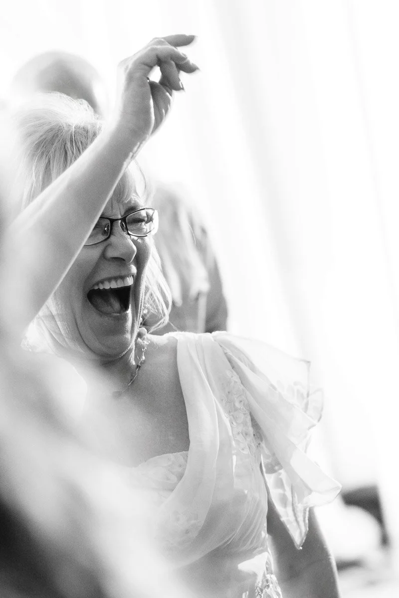 A joyful woman with glasses laughs heartily, her arm raised. She's wearing a light, flowy dress. The black and white setting conveys a lively, celebratory mood.