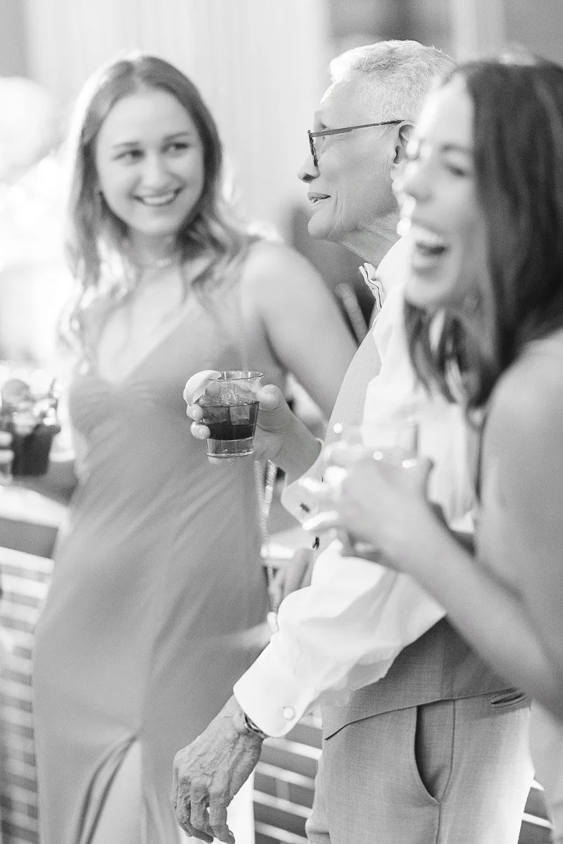 A black and white photo of three people at a party, laughing and holding drinks. They are casually dressed, conveying happiness and camaraderie.