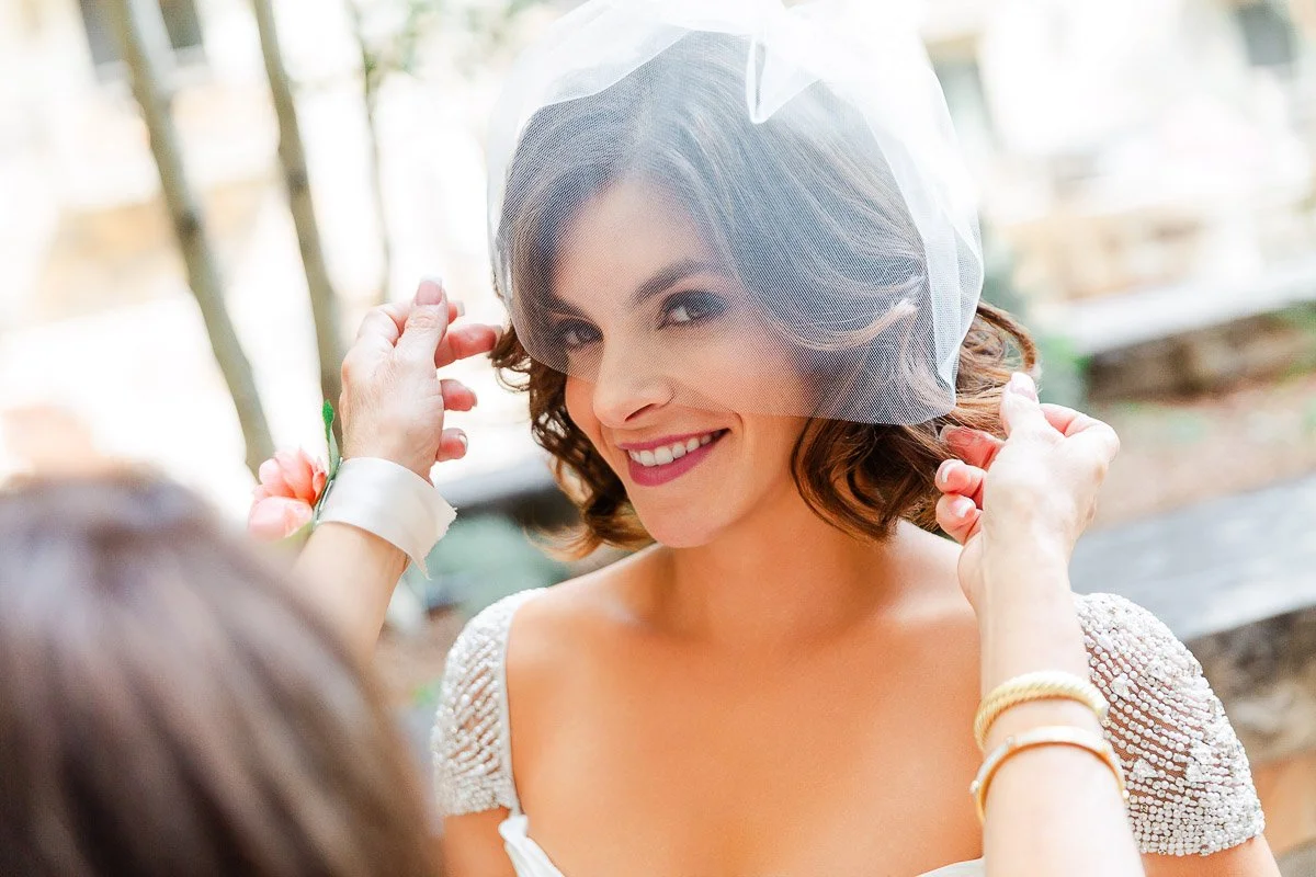 Smiling bride with short wavy hair wearing a veil, as two hands adjust it. She exudes joy and anticipation, outdoors in soft lighting.