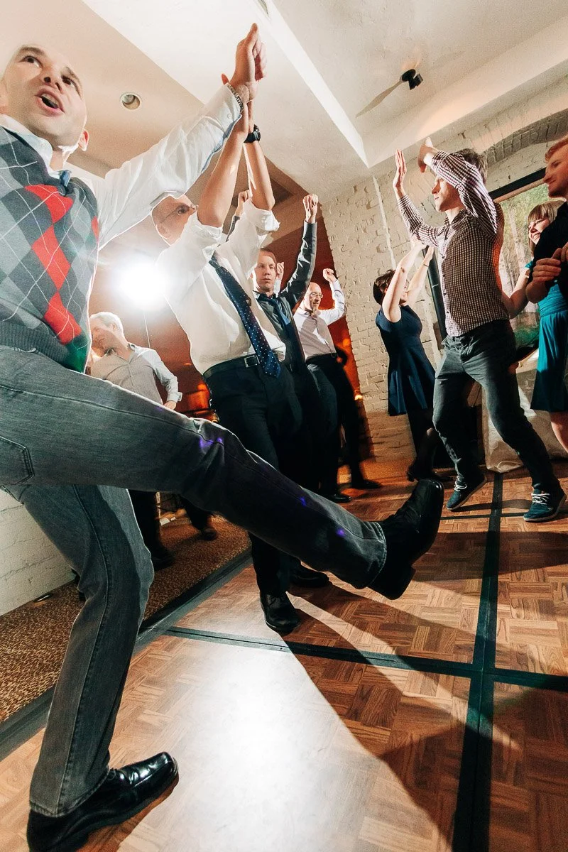 A group of people energetically dancing on a wooden floor, raising their hands and legs. The scene is lively, filled with joyful expressions.