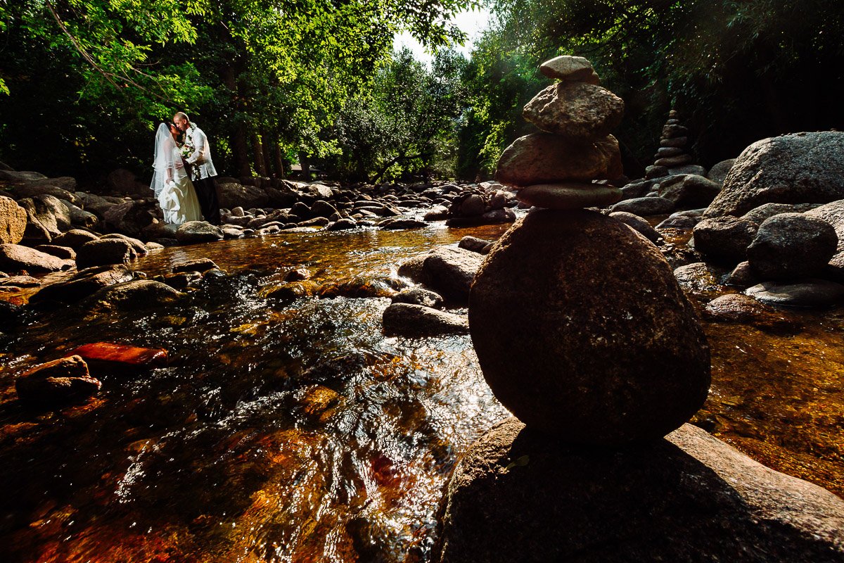 A couple in wedding attire embraces by a serene, Boulder Creek surrounded by lush greenery. Foreground features a balanced stone stack, creating a peaceful, romantic scene captured by Boulder Wedding Photographer tomKphoto