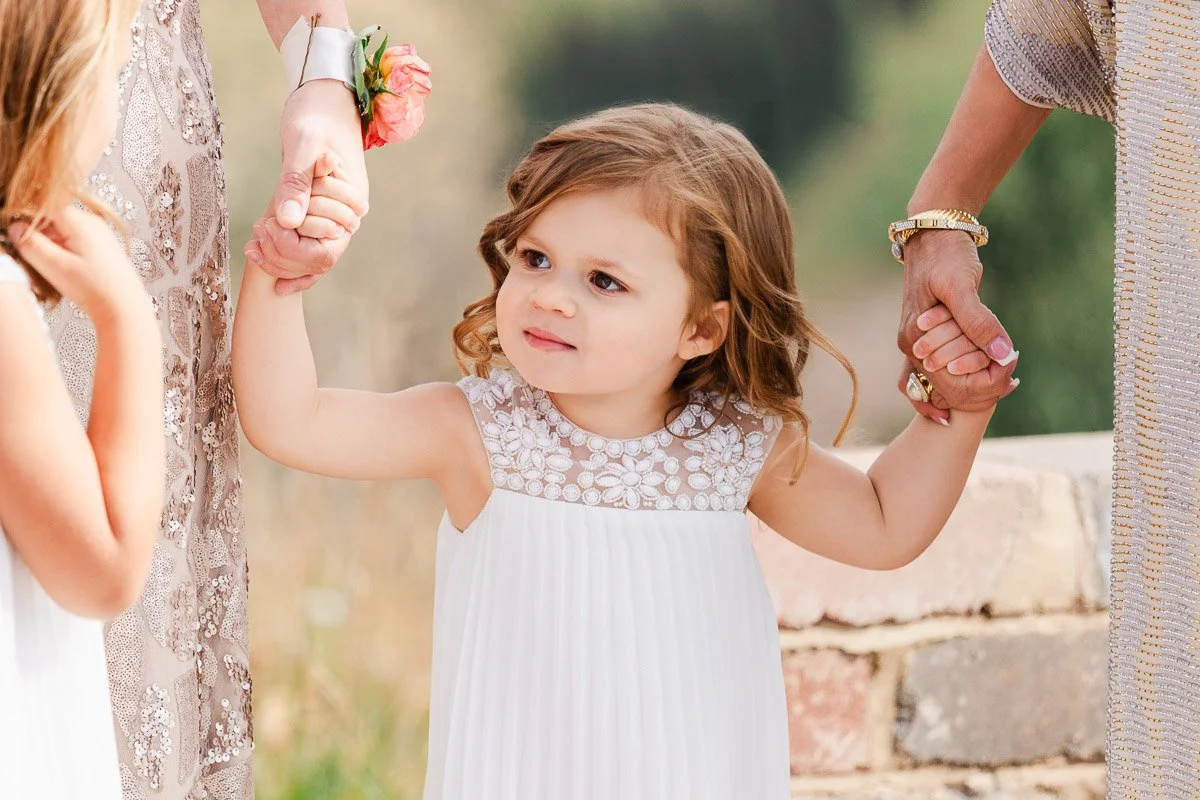 A young girl in a white dress with floral details holds hands with two adults, expressing a joyful and serene mood. A natural, blurred backdrop sets a warm tone.