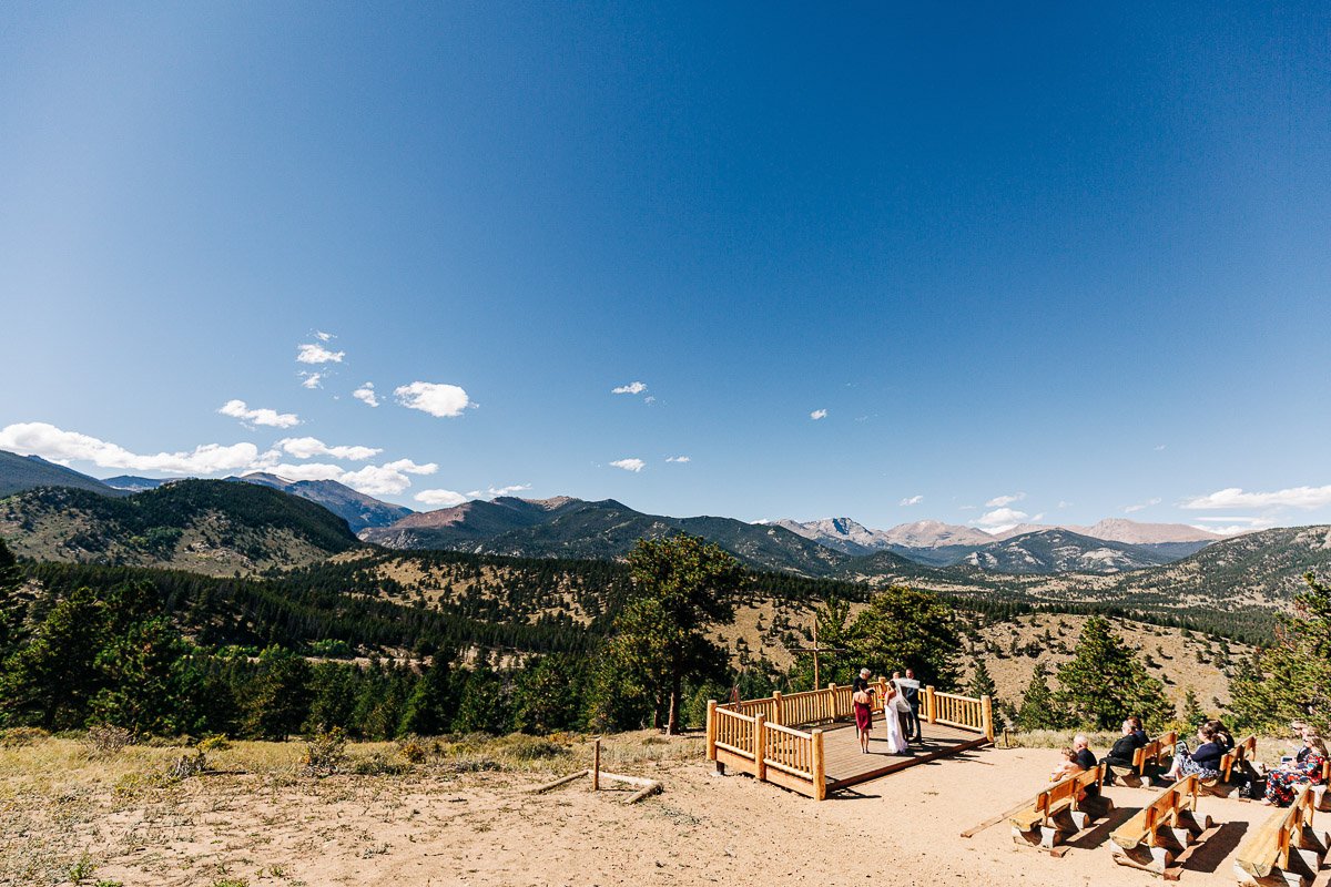 Outdoor wedding ceremony with guests seated on wooden benches, surrounded by a vast mountainous landscape under a clear blue sky. Peaceful and scenic, captured by YMCA of the Rockies wedding photographer tomKphoto