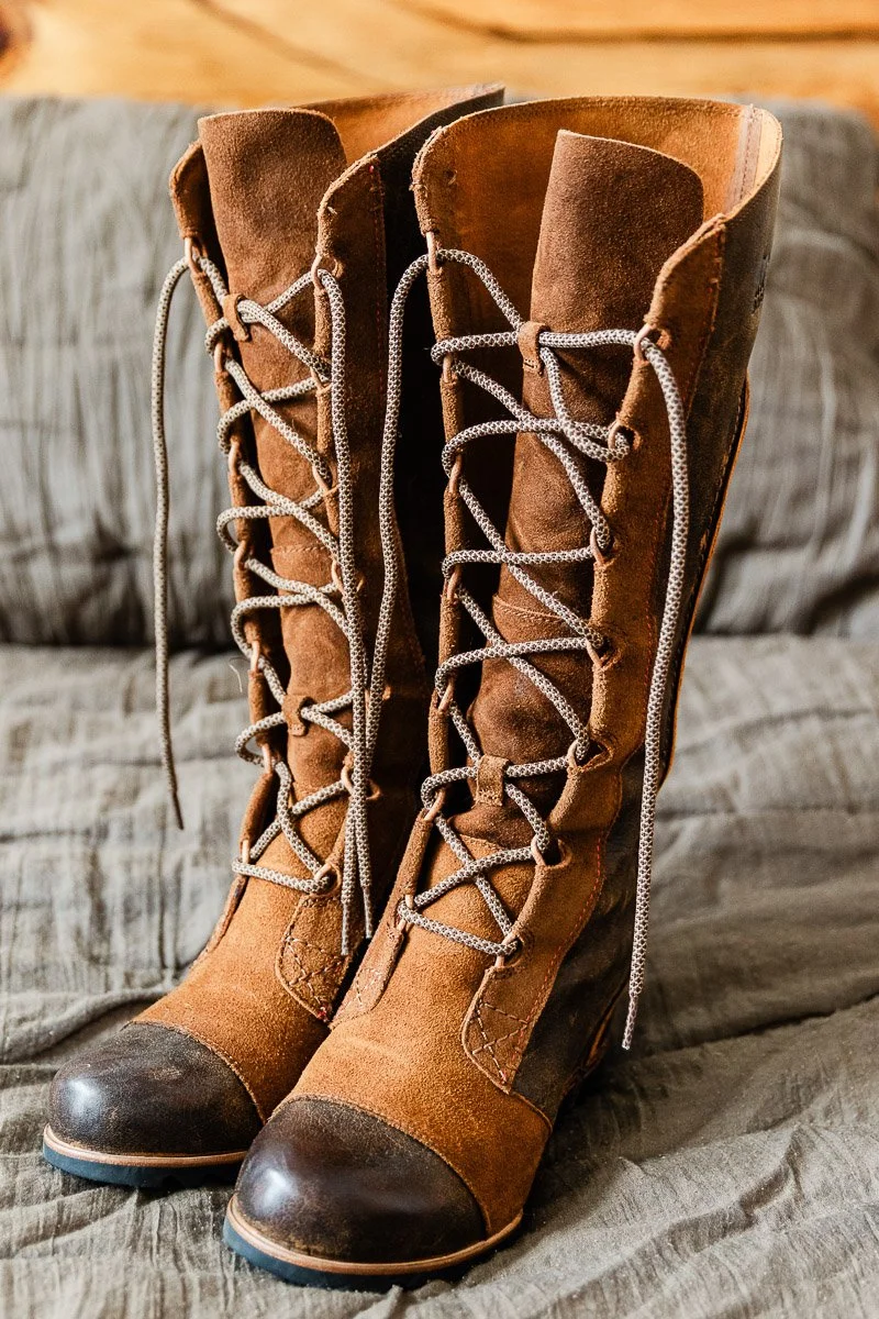 A pair of vintage brown leather boots with tall shafts and lace-up fronts sit on a textured gray fabric. The boots have dark brown toe caps.