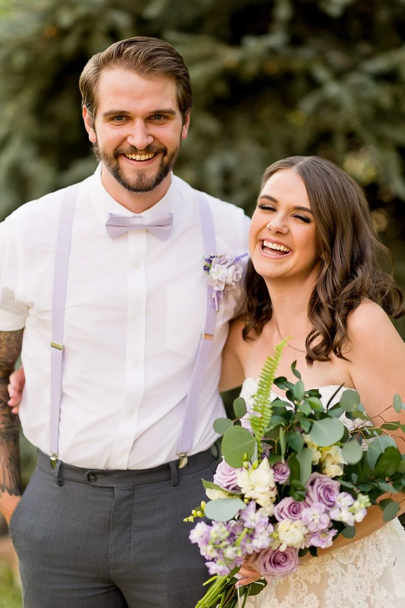 A joyful couple on their wedding day. The groom wears a white shirt with suspenders and lavender accents. The bride, in a lace dress, holds a bouquet of purple and white flowers.