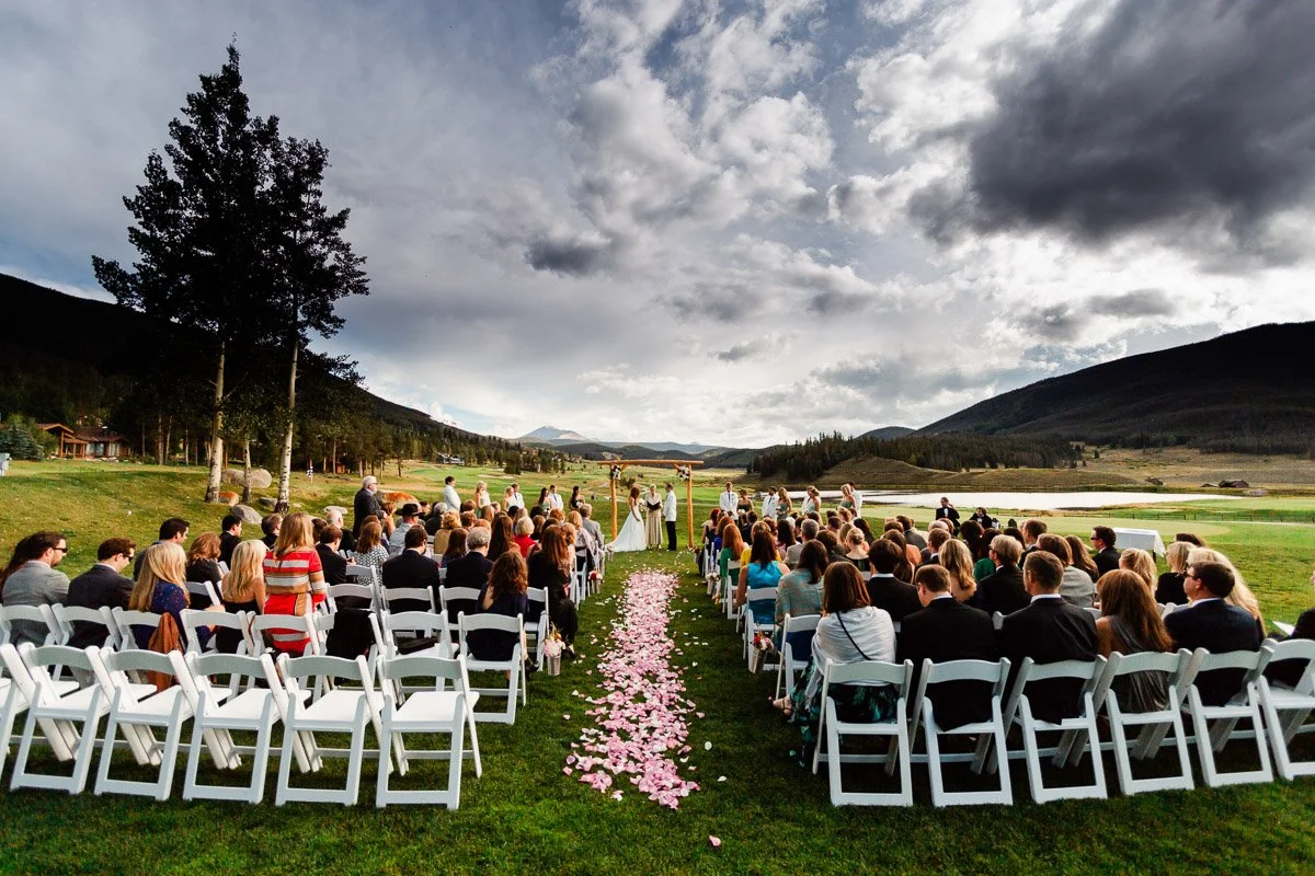 Outdoor Keystone Ranch wedding ceremony with guests seated on white chairs, facing a couple under an archway. Pink petals line the aisle. Overcast sky and scenic mountains surround.