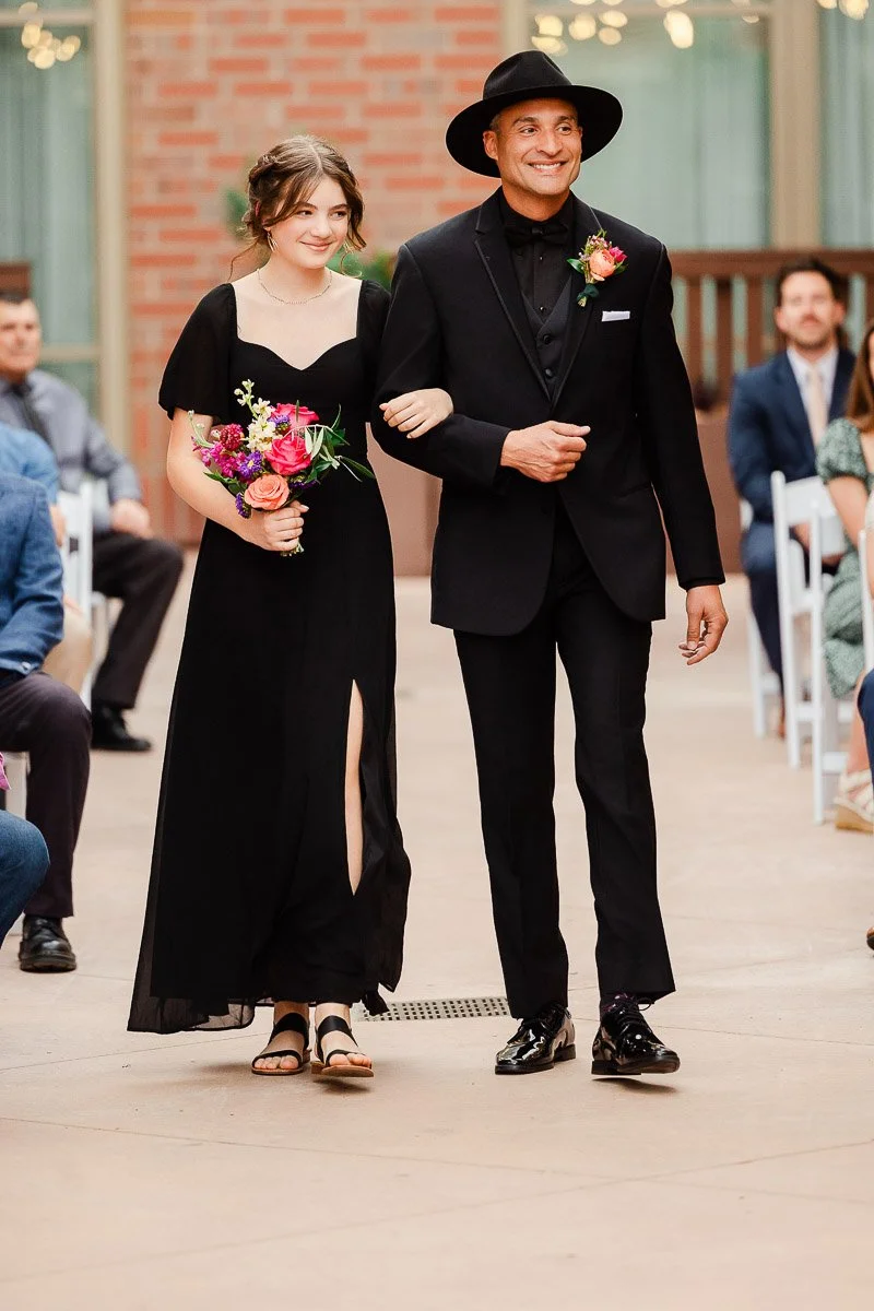 A young woman in a black dress and a man in a black suit with a hat walk arm-in-arm down an aisle. They hold bright flowers, smiling warmly.