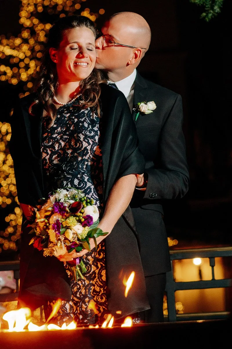 A smiling woman in a black lace dress holds a vibrant bouquet, while a man in a suit kisses her cheek tenderly. Warm lights twinkle softly in the background.