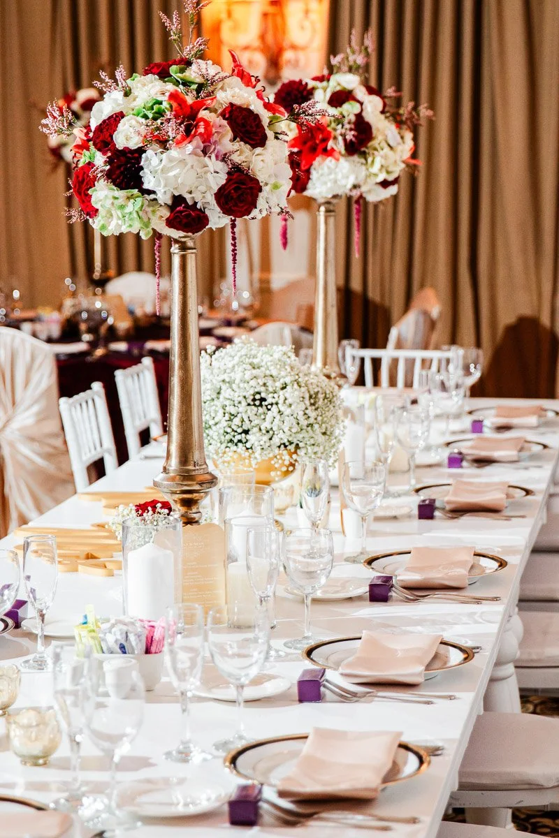 Elegant banquet table with tall floral centerpieces of red and white flowers. Set with fine china, glassware, and neutral napkins in a formal setting.