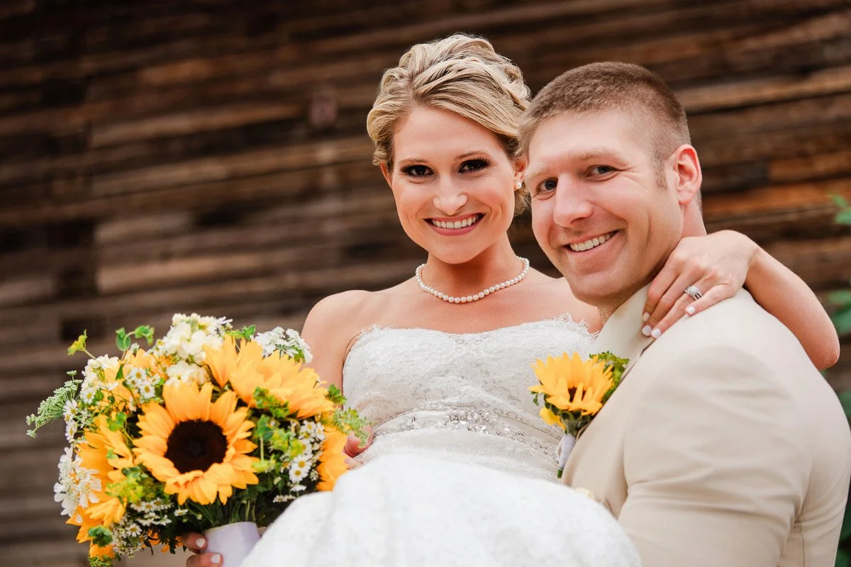 Bride in white dress smiles while being held by groom in beige suit. She holds a bouquet of sunflowers, with a rustic wooden backdrop. Joyful and romantic.