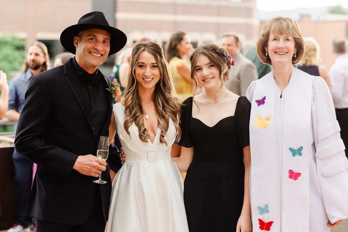 A group of four smiling people at an outdoor event. One man in a black suit and hat holds a champagne glass. Next to him, a woman in a white dress, another in a black dress, and a woman in a white dress with colorful butterfly appliqués.