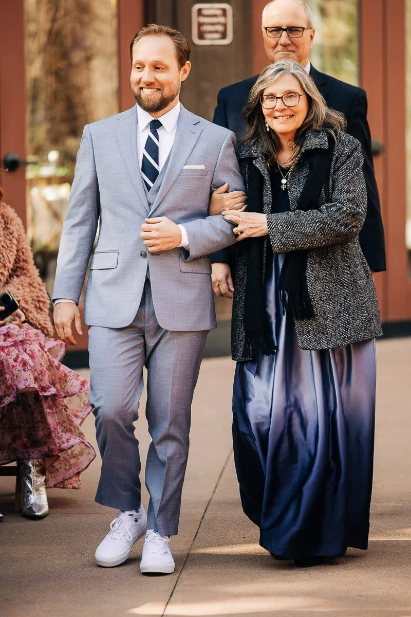 A smiling man in a light gray suit and white sneakers walks arm-in-arm with a woman in a flowing blue dress and gray coat, conveying joy and elegance.