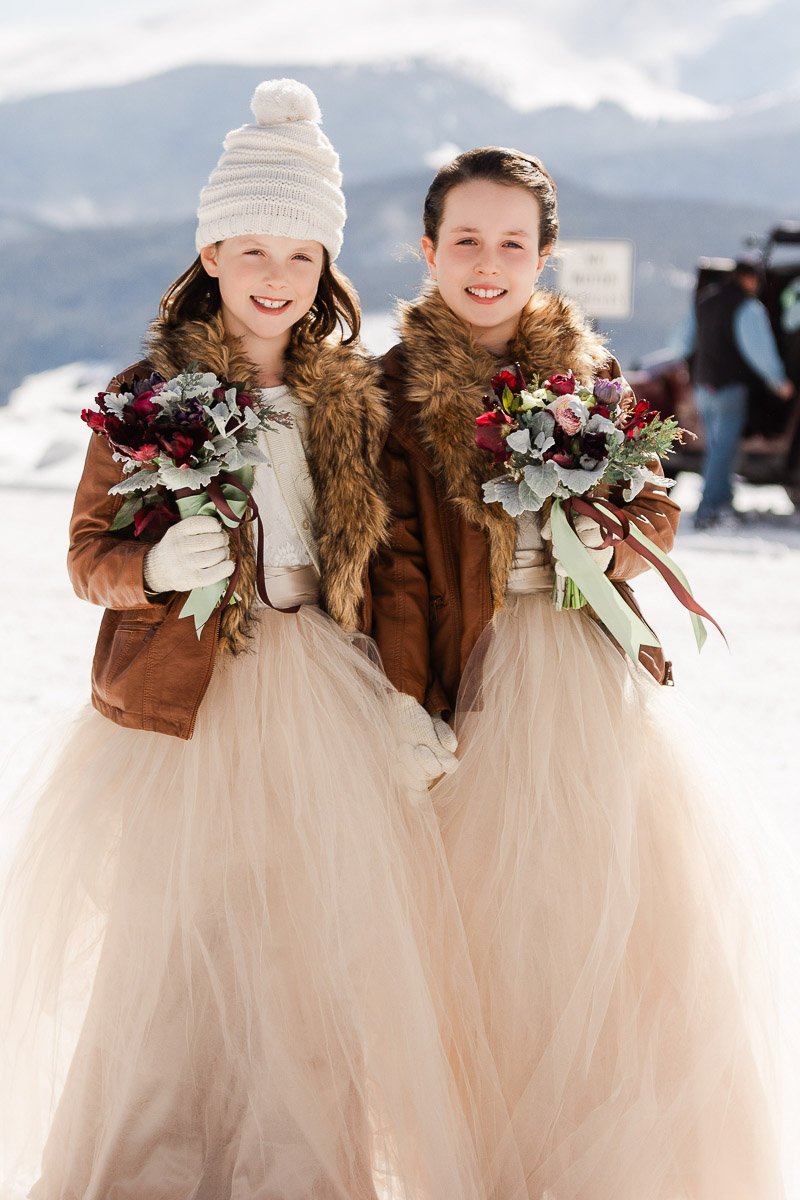 Two smiling girls in brown jackets with fur collars and peach tulle skirts stand in a snowy landscape, holding floral bouquets, exuding a joyful, winter vibe.
