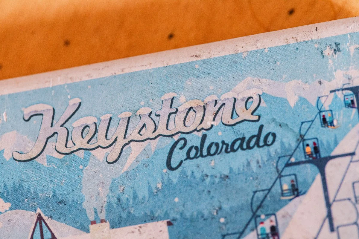 Vintage poster featuring "Keystone Colorado" in serif font, with snowy mountains, pine trees, and a ski lift against a faded blue background.