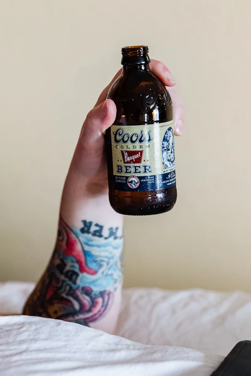 A tattooed hand holds a brown bottle of Coors Banquet Beer against a neutral background. The scene conveys a casual, relaxed mood.