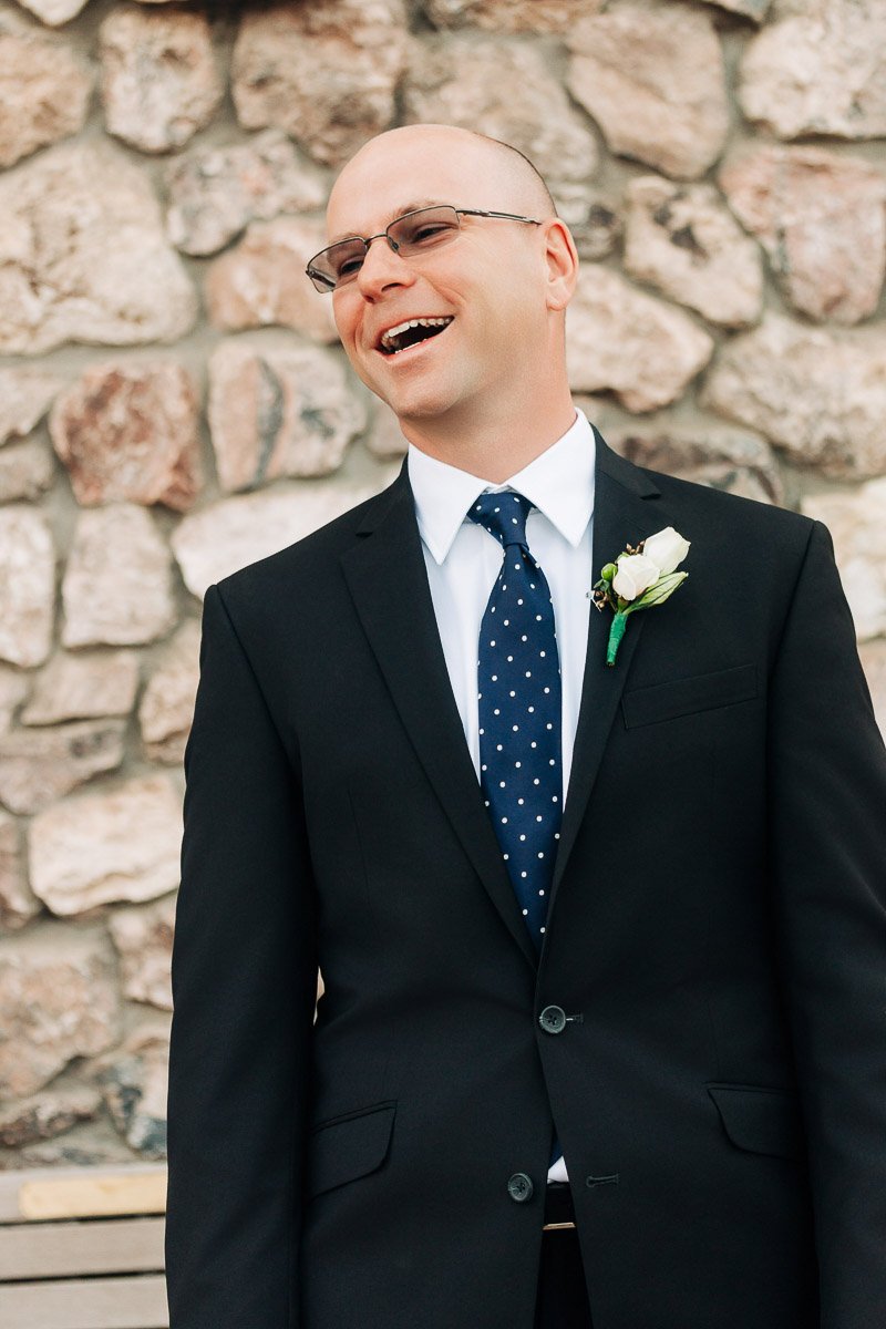 Smiling man in a black suit with a polka dot tie and white boutonniere stands against a stone wall, conveying joy and elegance.