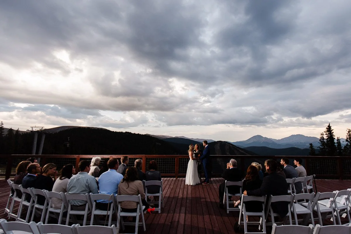 A couple stands at an outdoor wedding ceremony on a wooden deck with a mountainous backdrop. Guests sit on white chairs under a dramatic cloudy sky.