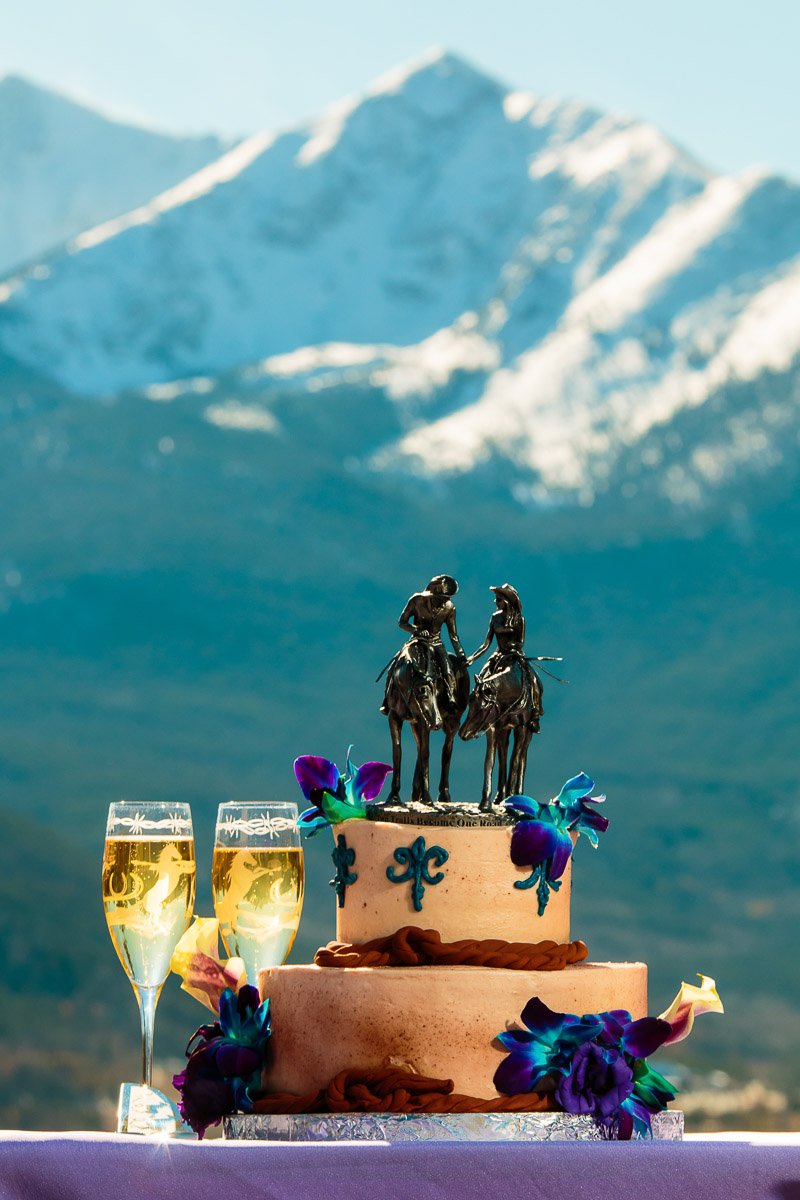 A two-tiered wedding cake with horse and rider figurines, adorned with blue and purple flowers, sits against a backdrop of snow-capped mountains. Two filled champagne glasses are beside it during a Sapphire Point wedding in Summit County, Colorado.