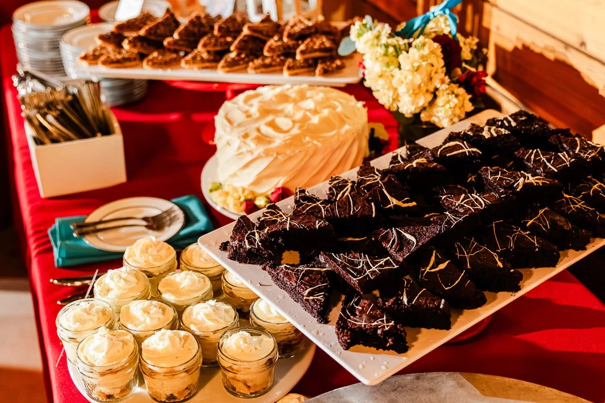 A vibrant dessert table with chocolate brownies, lemon mousse in jars, a white frosted cake, and pecan pastries on a red tablecloth, evoking indulgence.