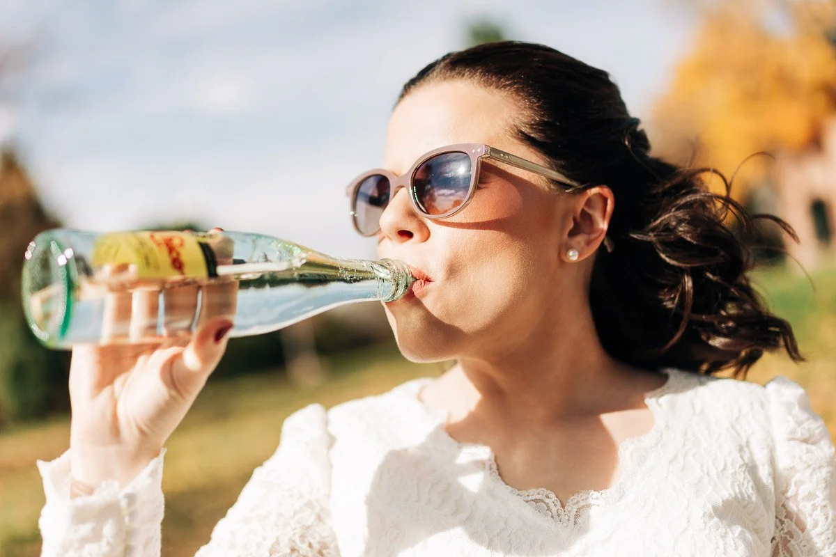Woman in sunglasses and a white top drinks from a glass bottle outdoors at a Wellshire Event Center wedding. Sunlight casts a warm glow, and autumn leaves are visible in the background.