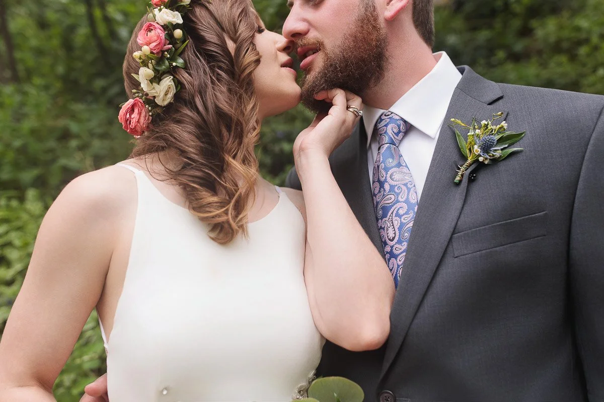 A bride and groom share an intimate moment outdoors. The bride wears a floral crown and white dress, while the groom is in a suit with a floral boutonniere.
