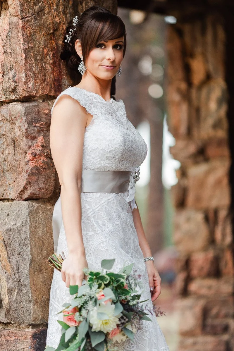 Bride in elegant lace gown stands beside rustic stone wall during a Boettcher Mansion wedding, holding a bouquet with greenery and peach roses. Her expression is serene and poised.