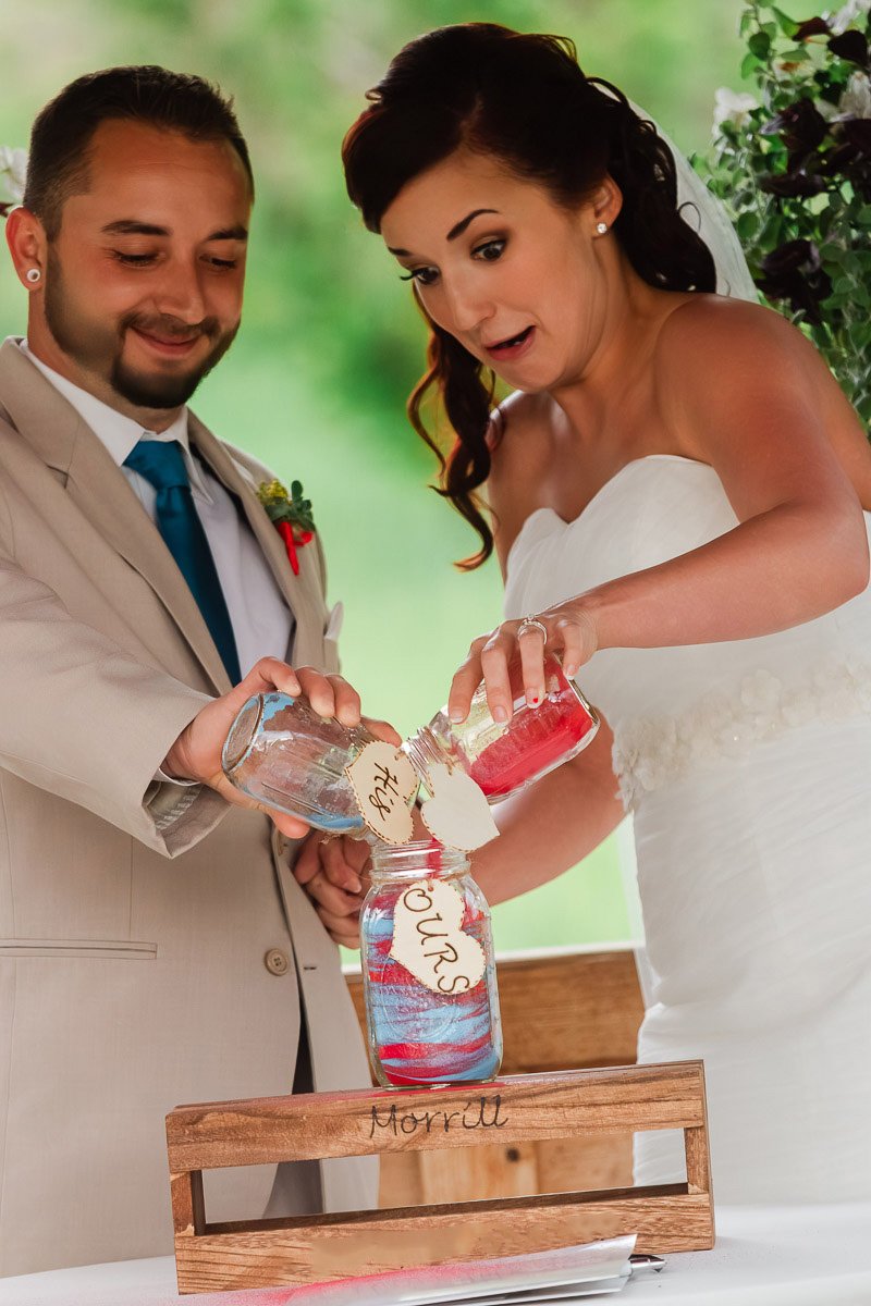 Bride and groom pour colored sand into a jar labeled "Ours," symbolizing unity. The bride looks surprised; the setting is lively and joyful.