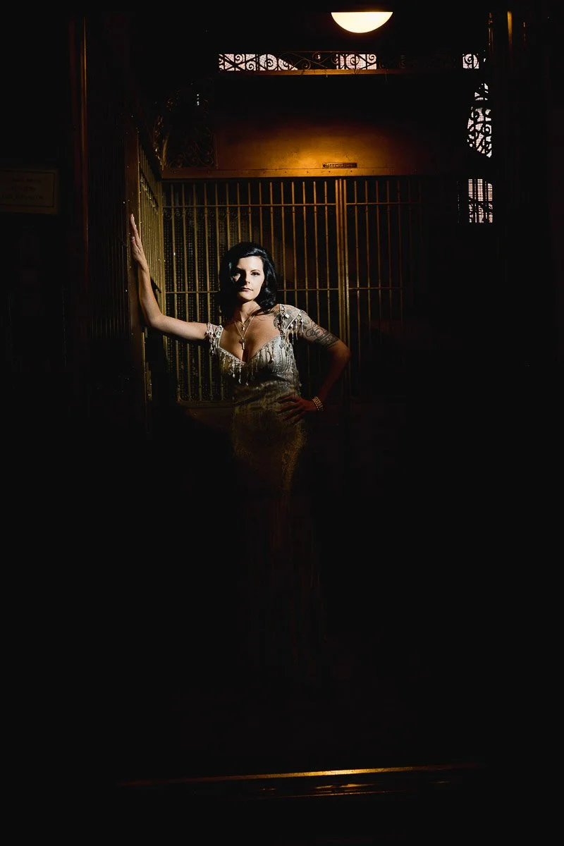 A woman in an elegant, shimmering gown poses confidently in a dimly lit vintage elevator. The mood is mysterious and glamorous during a Mining Exchange Hotel wedding in Colorado Springs, Colorado