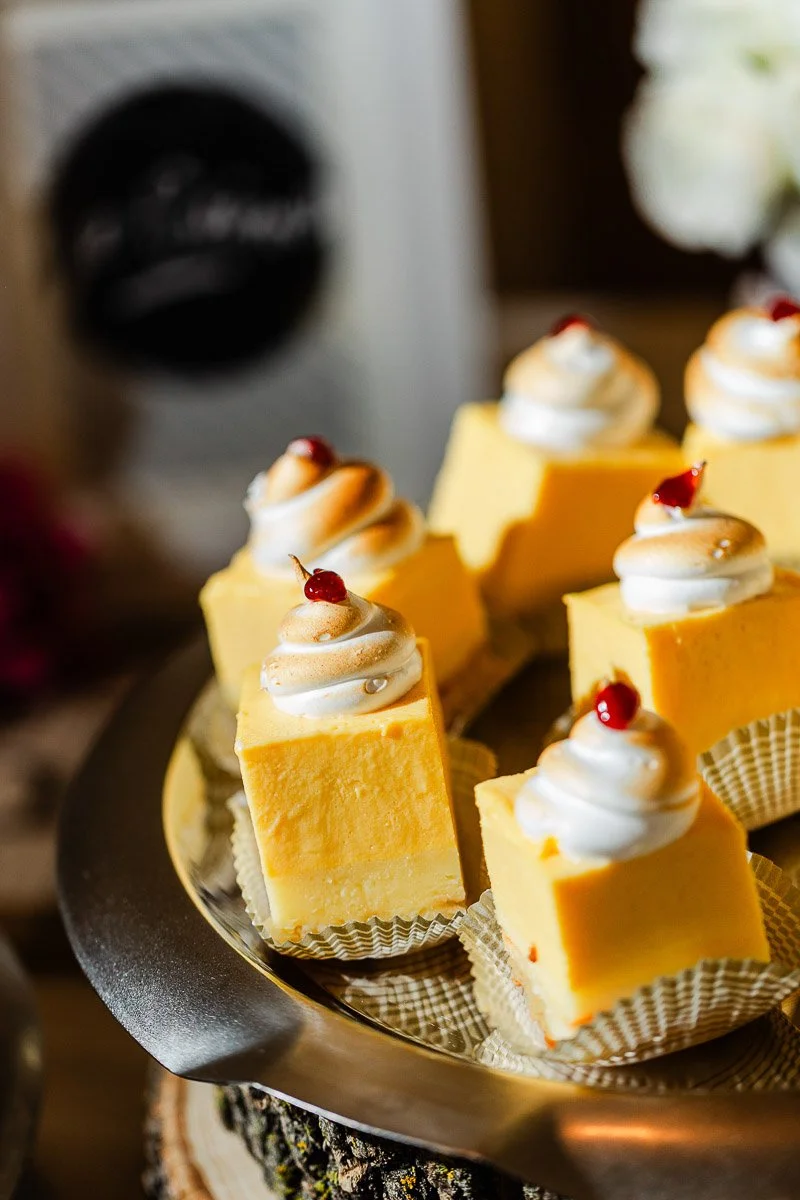 Slices of yellow square cake with dollops of whipped cream and small red cherries on top, elegantly arranged on a metal tray, creating a festive vibe.