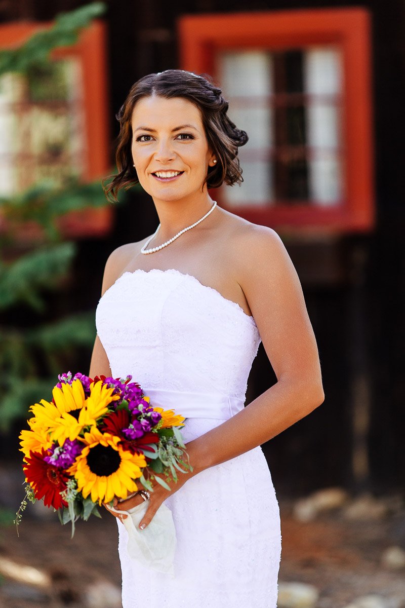 A bride in a strapless white dress smiles, holding a vibrant sunflower bouquet. She stands against a blurred rustic backdrop with red-framed windows captured by YMCA of the rockies wedding photographer tomKphoto