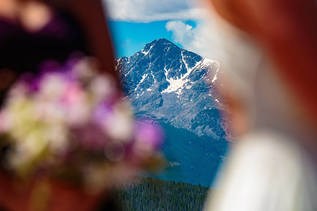 A majestic snow-capped mountain under a blue sky is framed by blurred figures and colorful flowers in the foreground, creating a dreamy, serene scene.